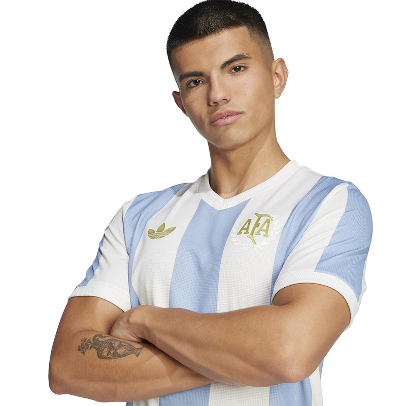 Escudo silkado dourado da Camisa Argentina 25/26 Adidas, destacando a parceria histórica.